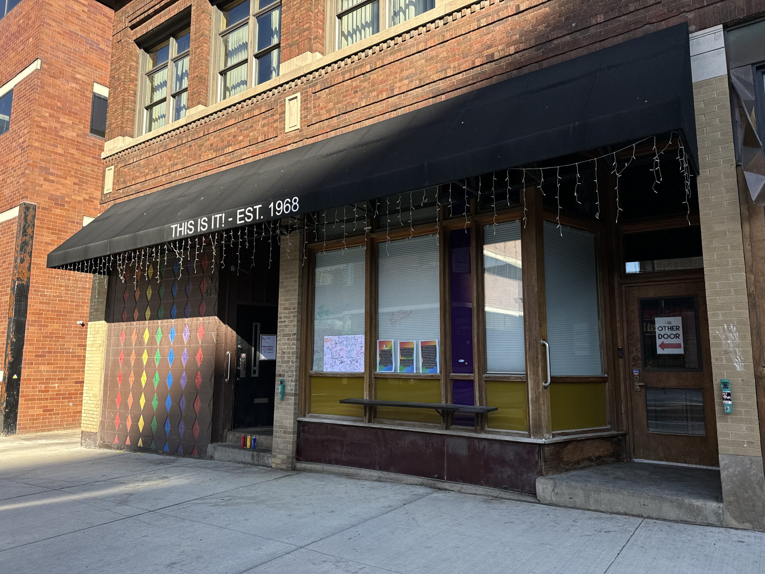 Entrance of a brick building with a black awning reading This Is It! - Est. 1968, colorful diamond patterns on the door, window displays, and a sign stating Use Other Door.