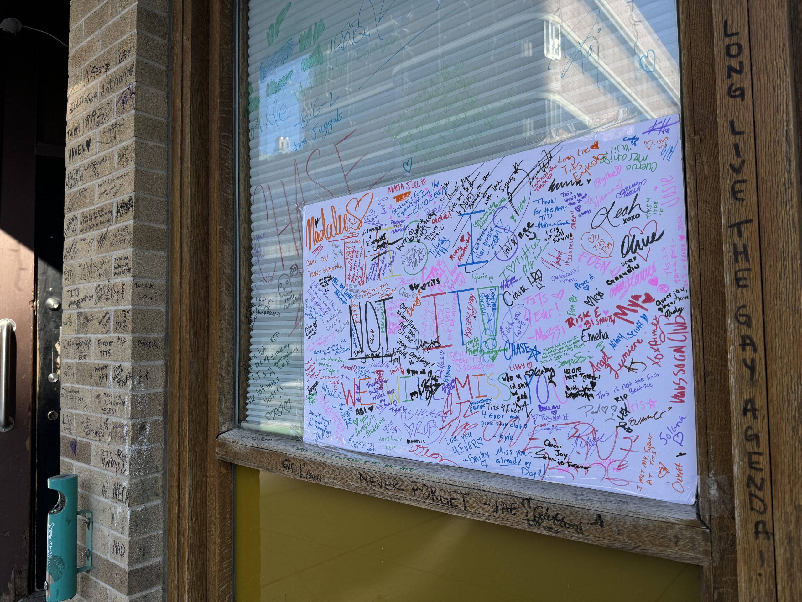 A window with colorful messages and signatures on a white paper. Brick walls and wooden frames are visible beside the glass.