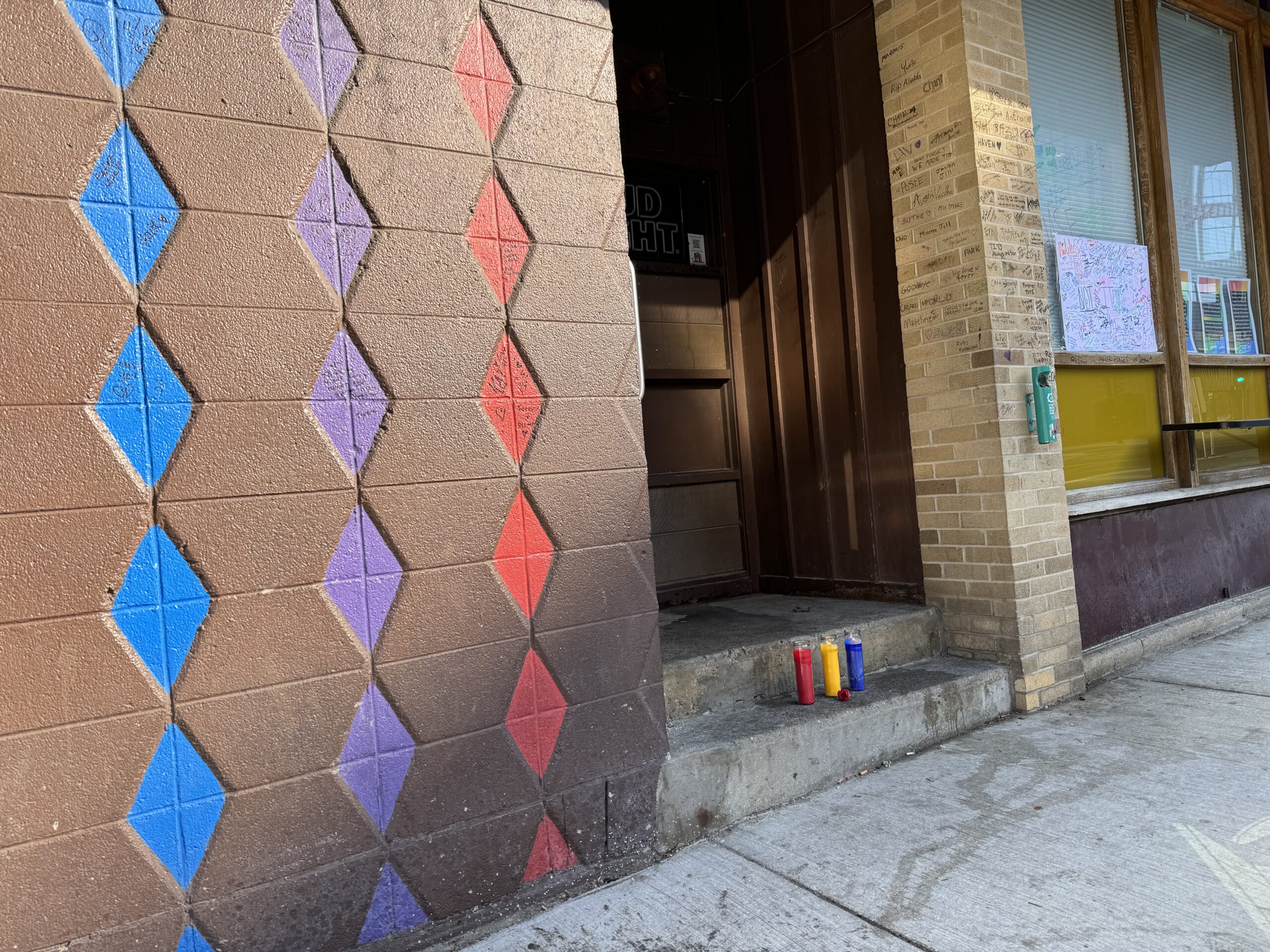 A wall with a diamond pattern in blue and red, next to a brick entrance with paint cans on the step. Nearby windows display papers.