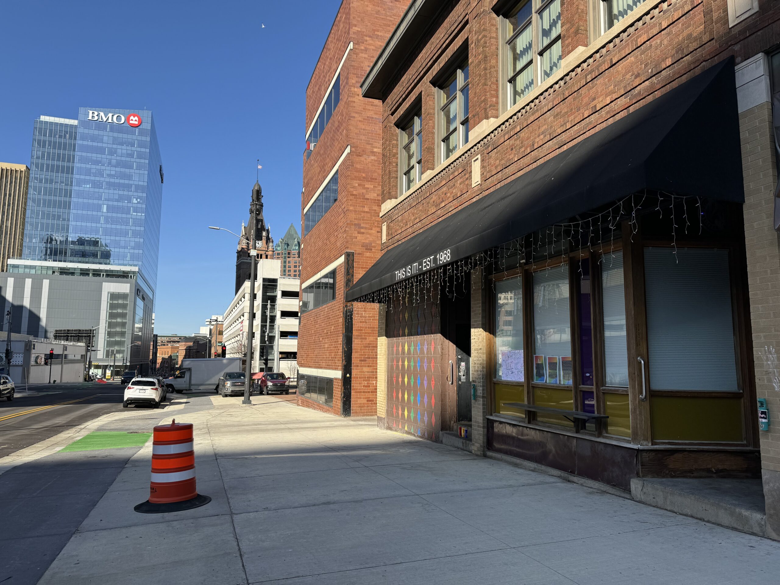 Urban street view with brick buildings, a sidewalk, and a traffic cone in the foreground. A tall modern building with a BMO logo is visible in the background under a clear blue sky.