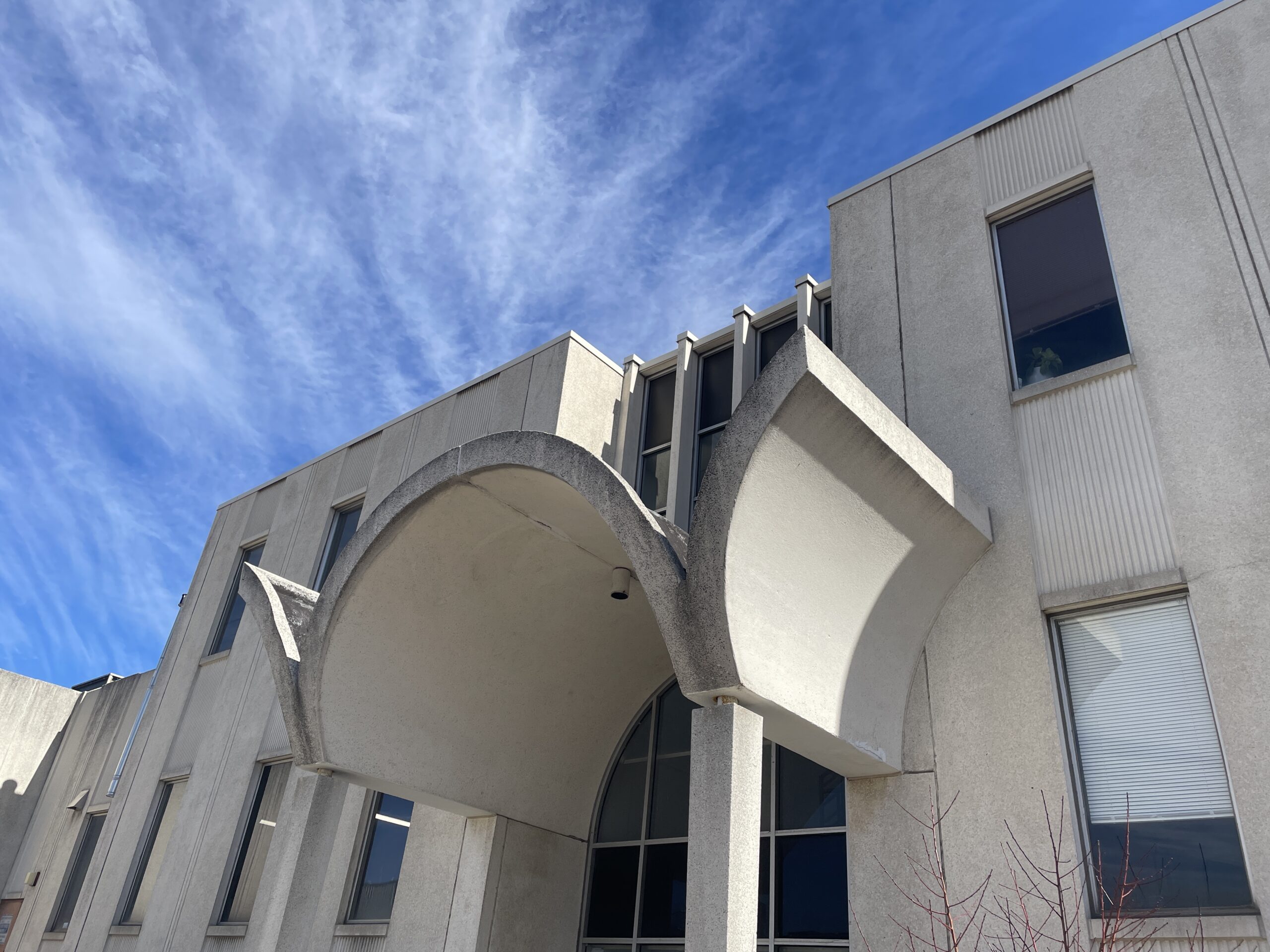 Concrete building with an arch detail at the entrance, vertical windows, and a clear blue sky with wispy clouds overhead.