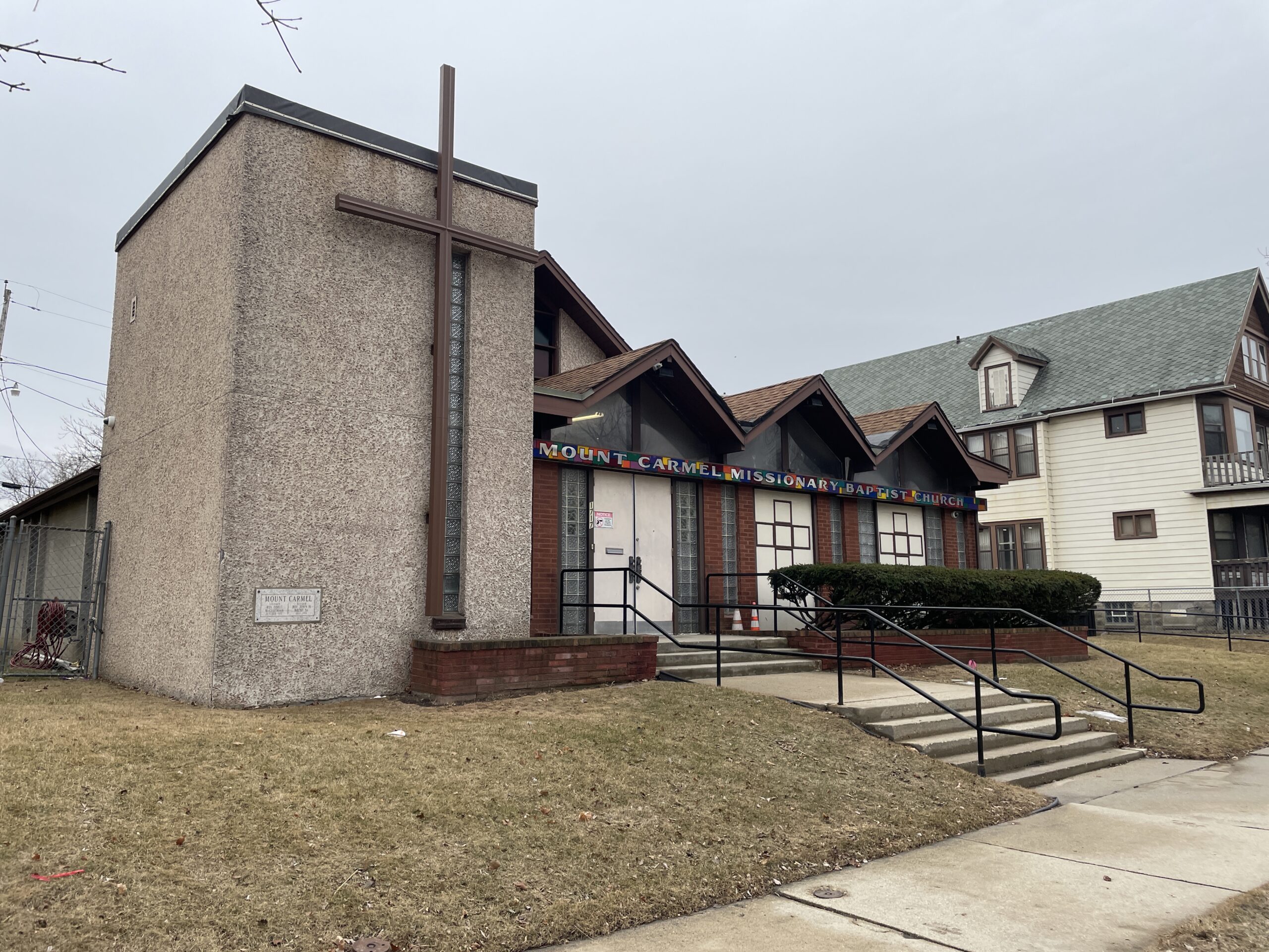 A church building with a large cross on a concrete tower, featuring a sign reading Mount Carmel Missionary Baptist Church, with stairs and a ramp leading to the entrance.