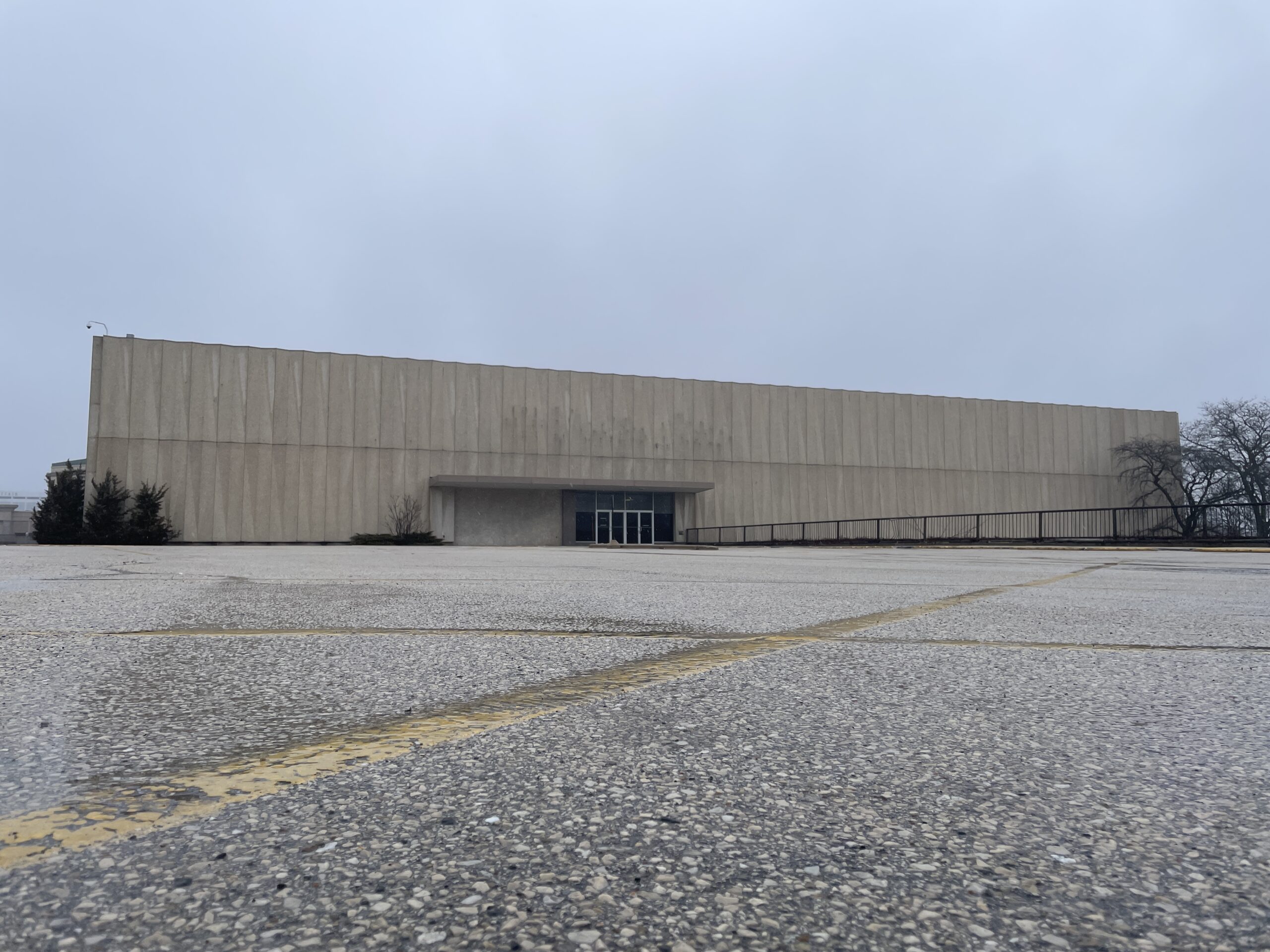 Large, empty parking lot in front of a beige, windowless building on a cloudy day. Sparse trees and bushes are visible on the sides.