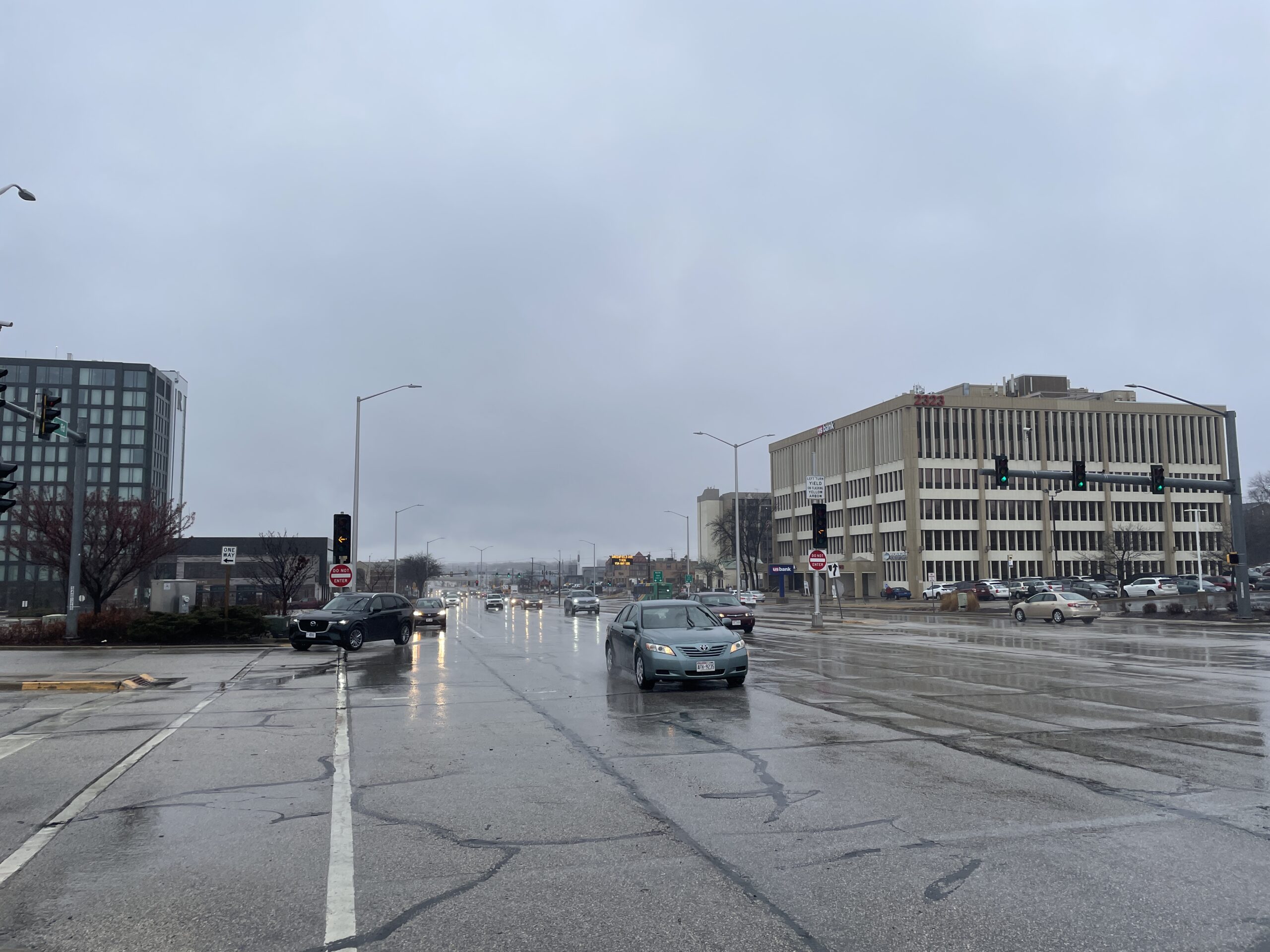 Cars drive on a wet, overcast urban street with traffic lights and office buildings, under a cloudy sky.