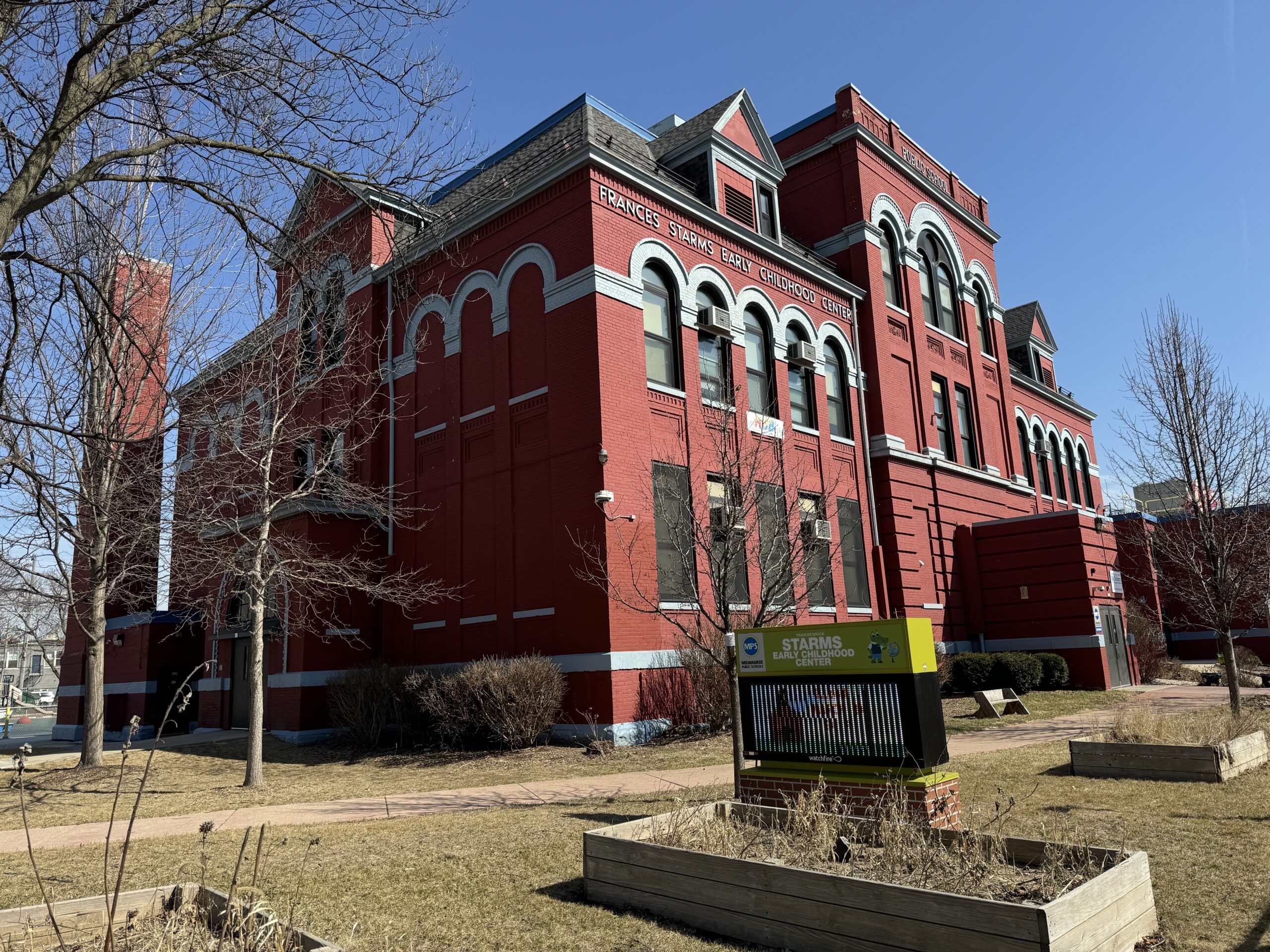 Red brick building with arched windows and a sign reading Francis Xavier Warde School. The foreground features leafless trees, bushes, and a sign about Stagg Field. Clear, sunny day.