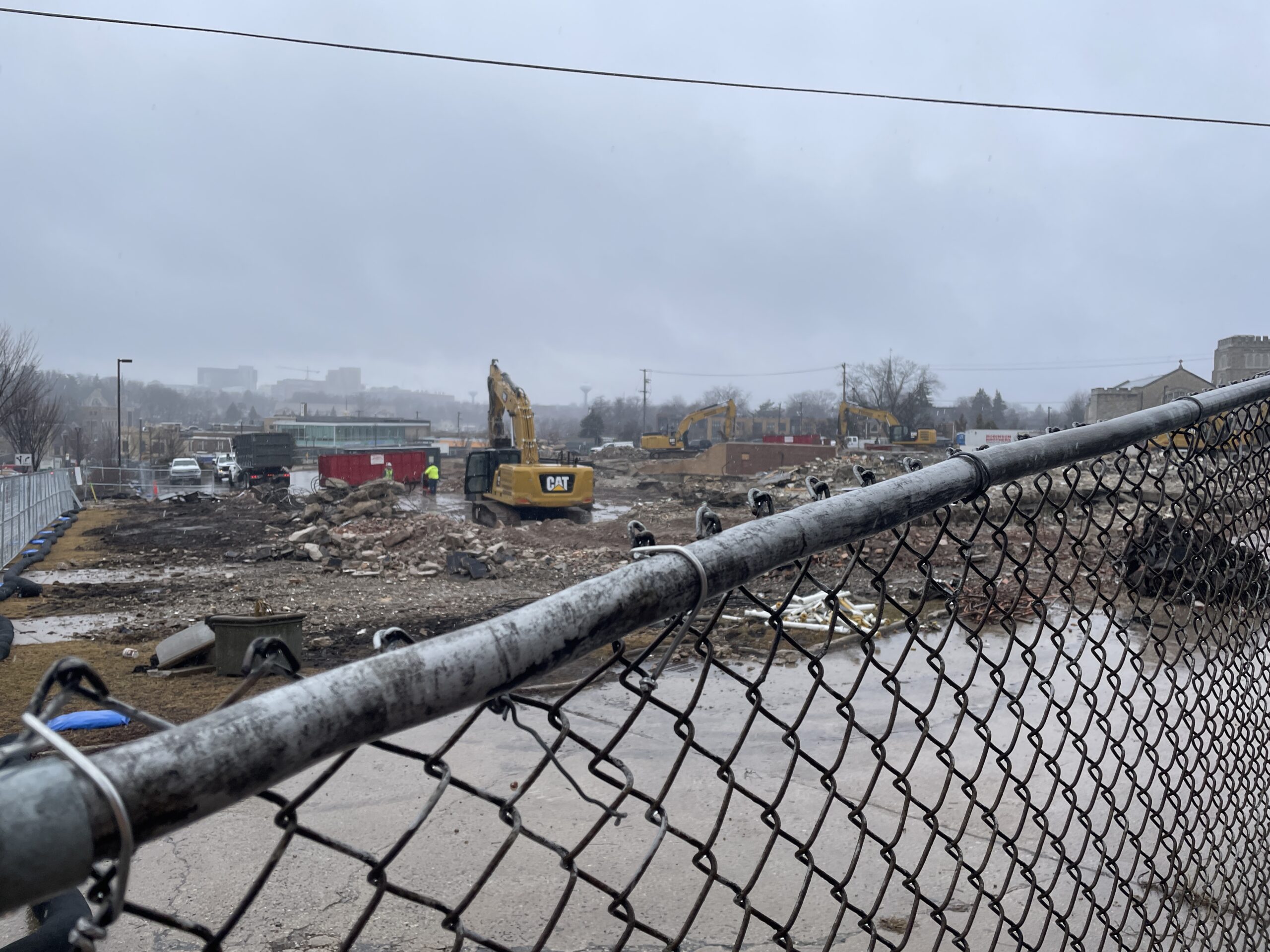 Construction site on a rainy day with machinery and debris visible behind a chain-link fence.