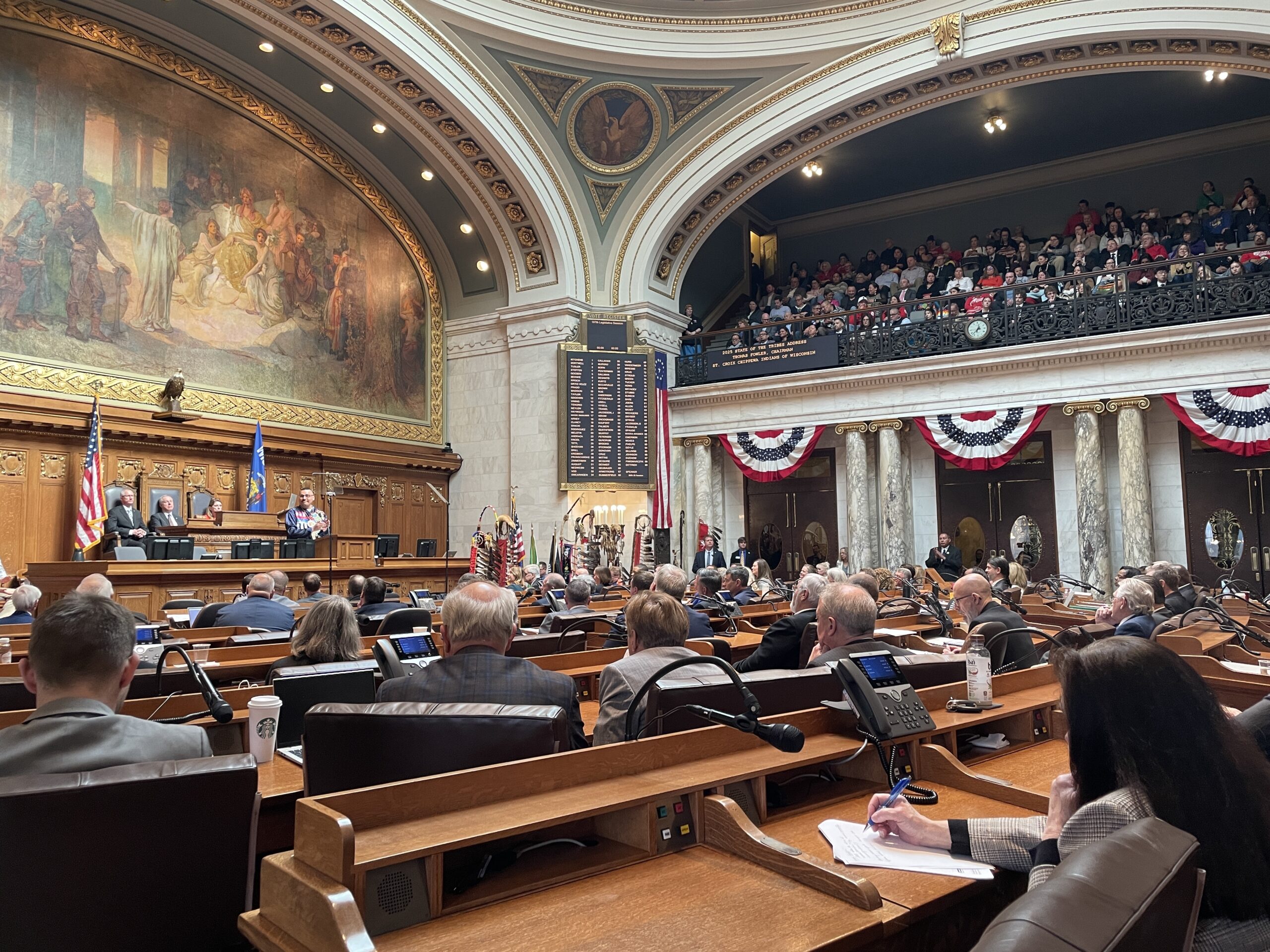 A legislative session in progress inside a large, grand chamber with officials seated at desks and an audience in the balcony. Flags and murals decorate the space.