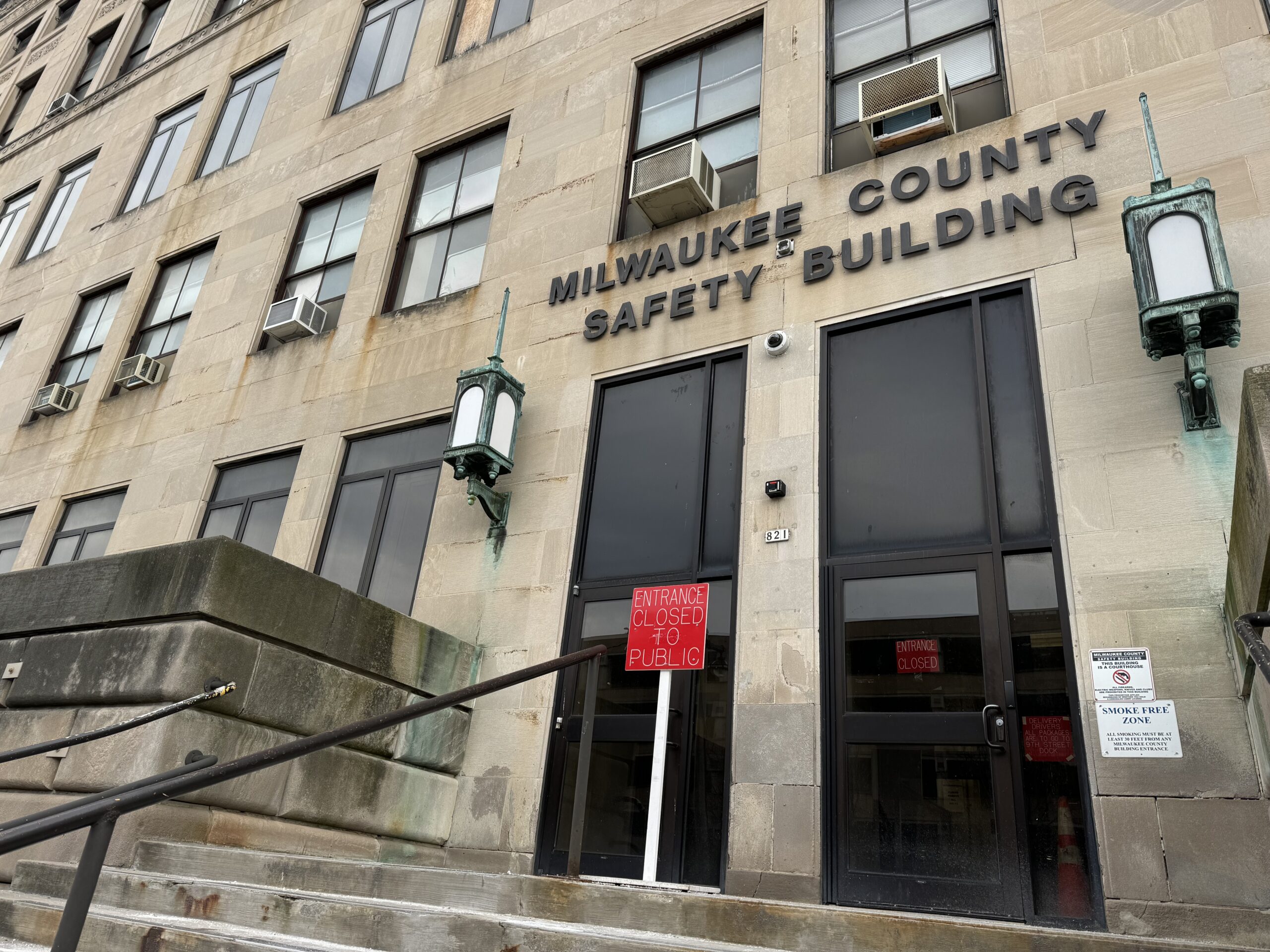 The image shows the entrance of the Milwaukee County Safety Building with signs indicating closure to the public. Steps and lanterns are visible near the entrance.