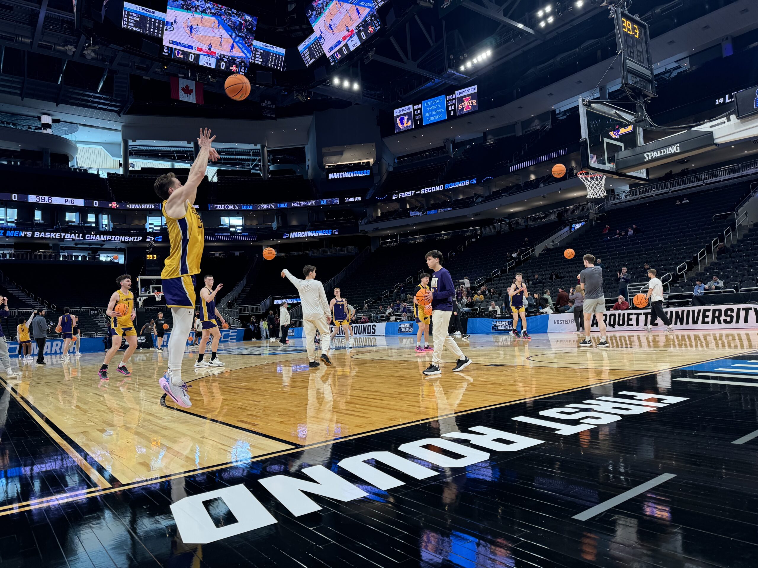 Basketball players practice on a court labeled First Round with a scoreboard and spectators in a large sports arena.