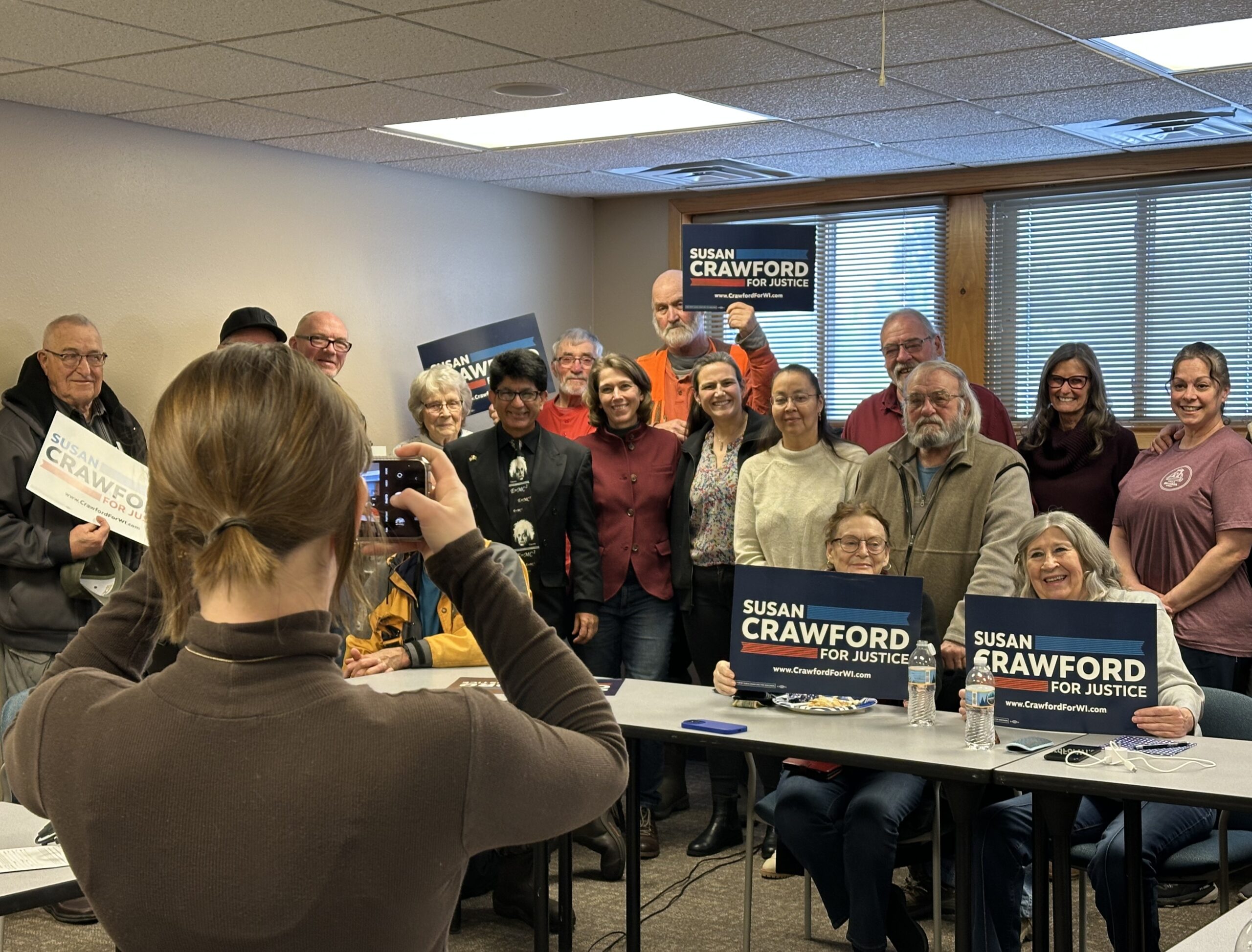 A group of people in a room, some holding Susan Crawford for Justice signs. A person in the foreground takes a photo of the group with a smartphone.