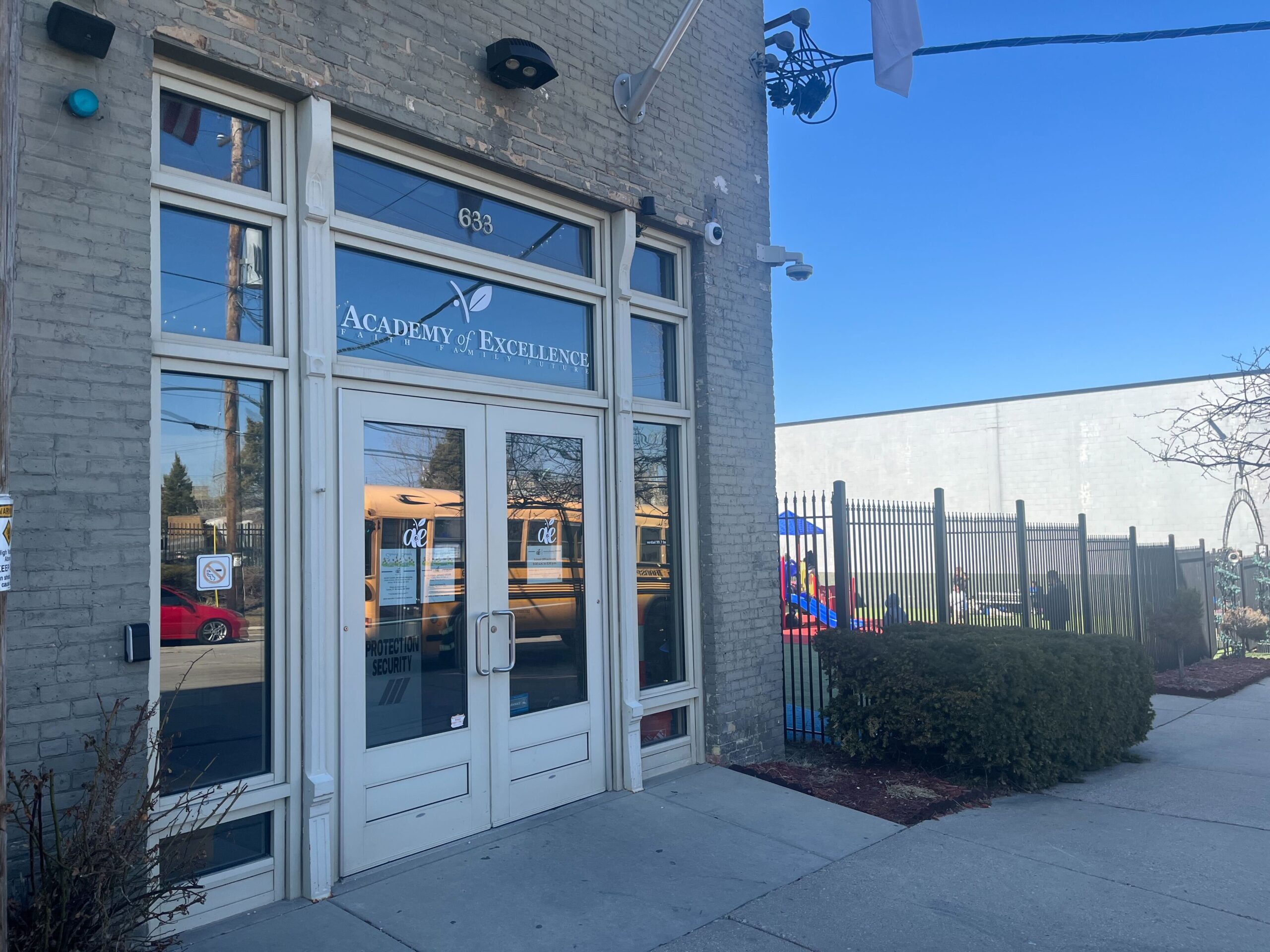 Entrance of a building labeled Academy of Excellence with large glass doors, a bus visible through windows, and a playground behind a fence to the right.