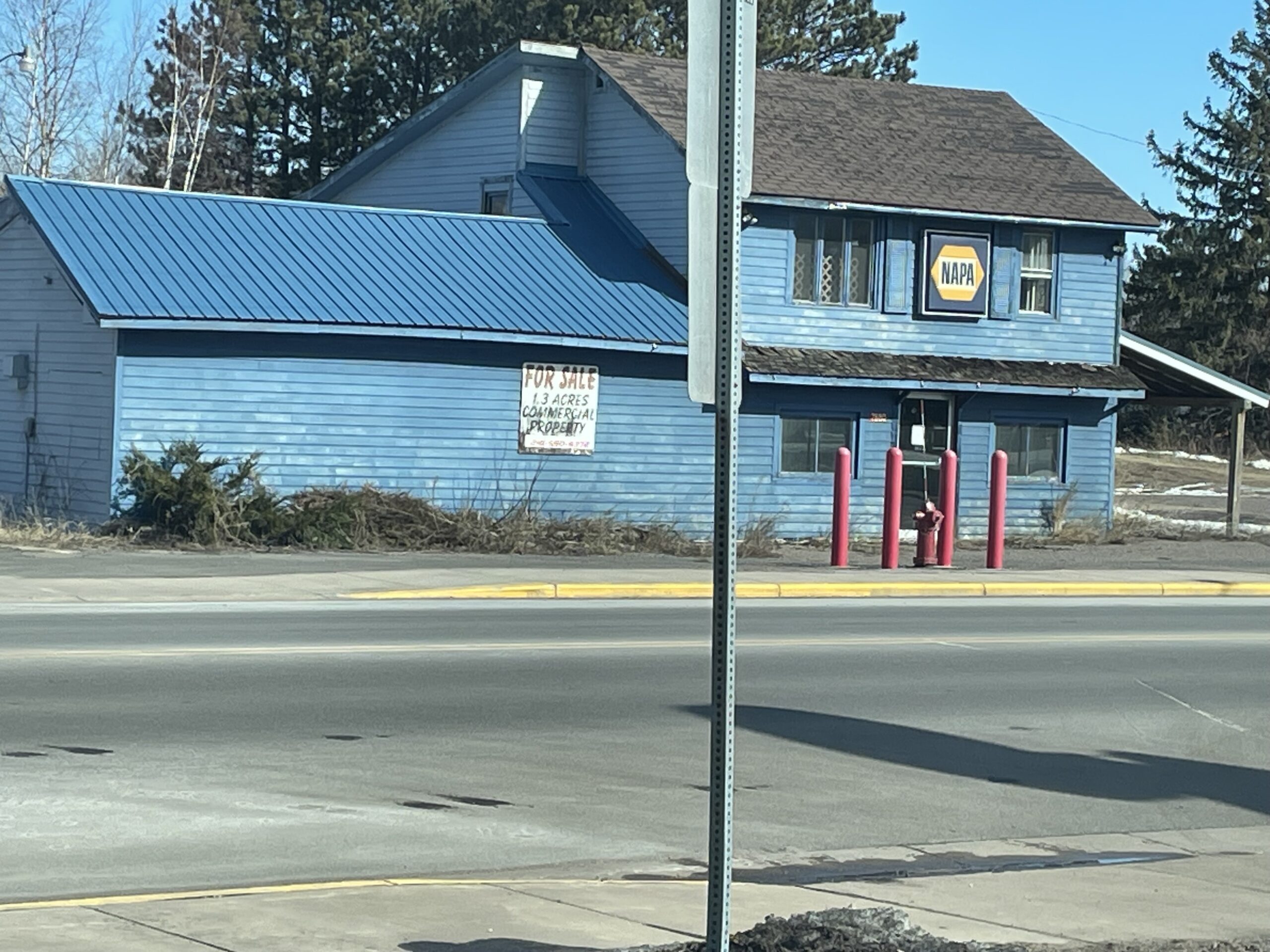 Blue building with a NAPA Auto Parts sign and a For Sale sign on the side, located near a road with a few trees in the background.