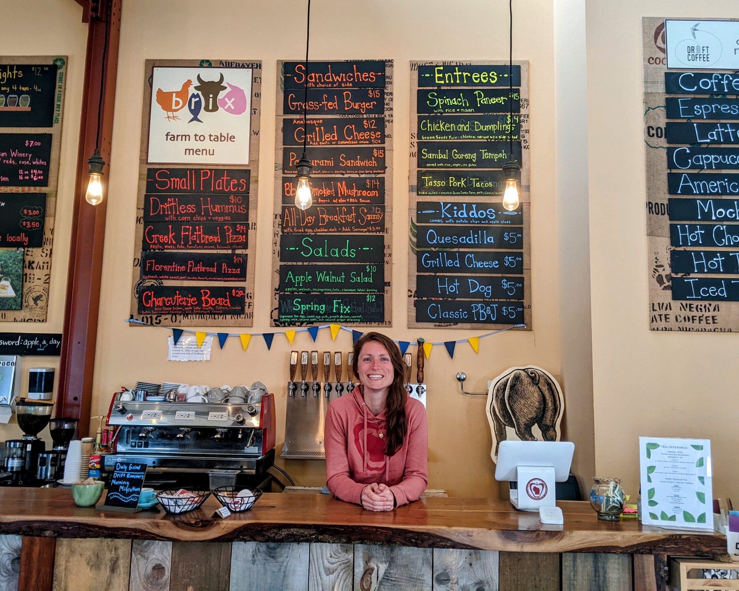 A woman stands behind a wooden counter at a cafe. Colorful menus on the wall display food and drink options. Coffee machine and register are visible on the counter.