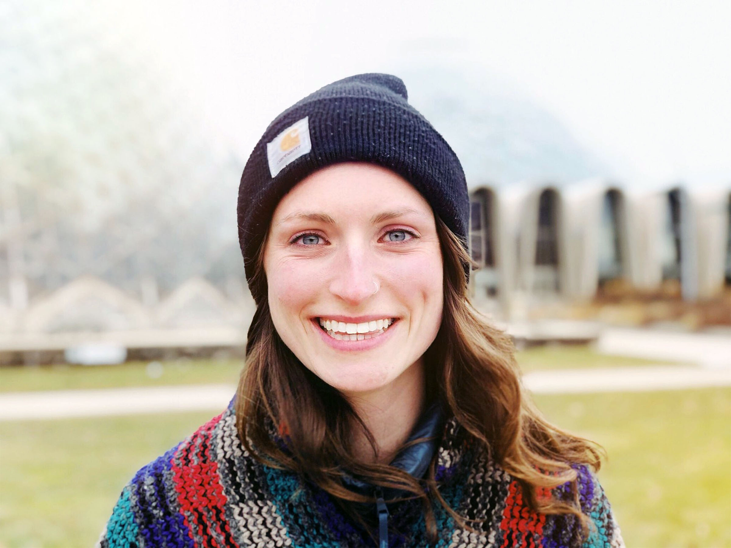 A woman in a black beanie and multicolored scarf smiles at the camera. Theres a blurred dome structure in the background.