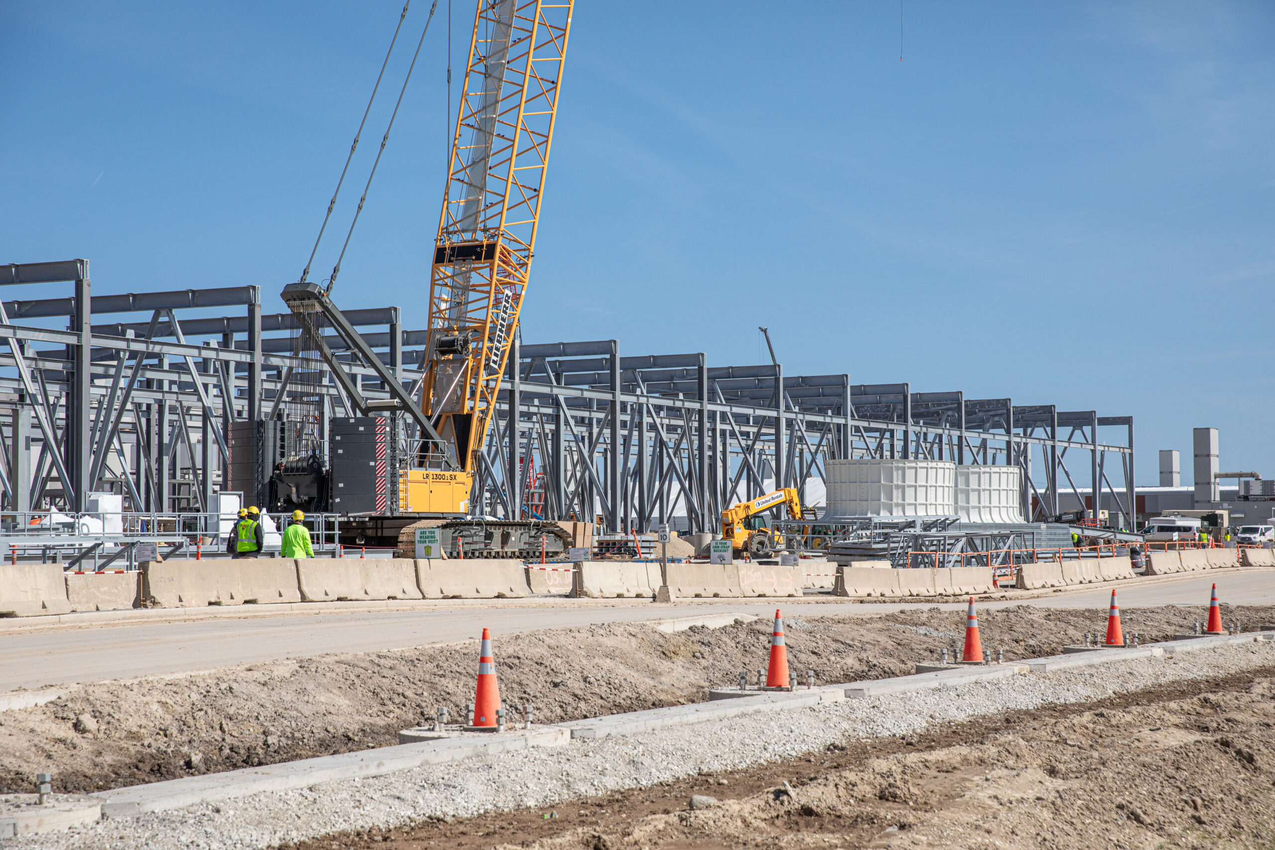 Construction site with large steel framework, a crane, and several workers in safety vests. Orange traffic cones line the foreground, and the sky is clear.