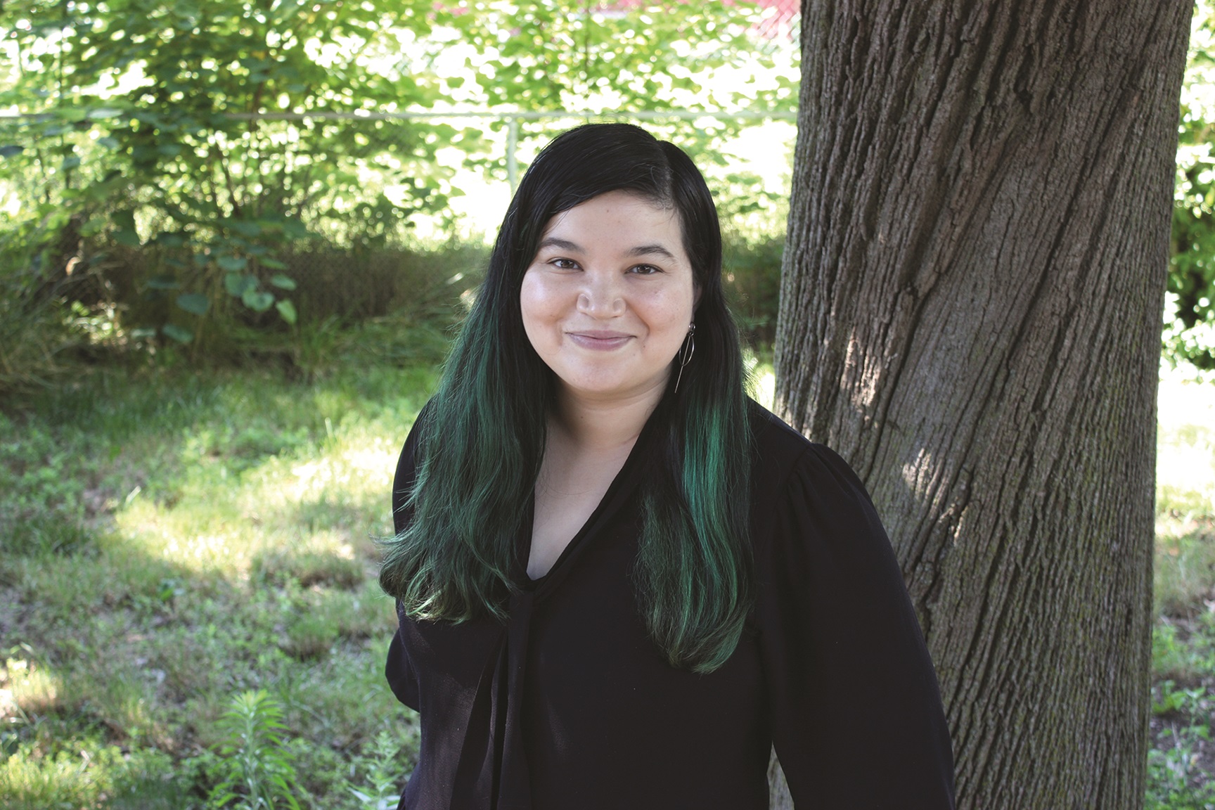 Person with long dark hair, partially dyed green, standing outdoors by a tree, wearing a black top, smiling at the camera. Lush greenery in the background.