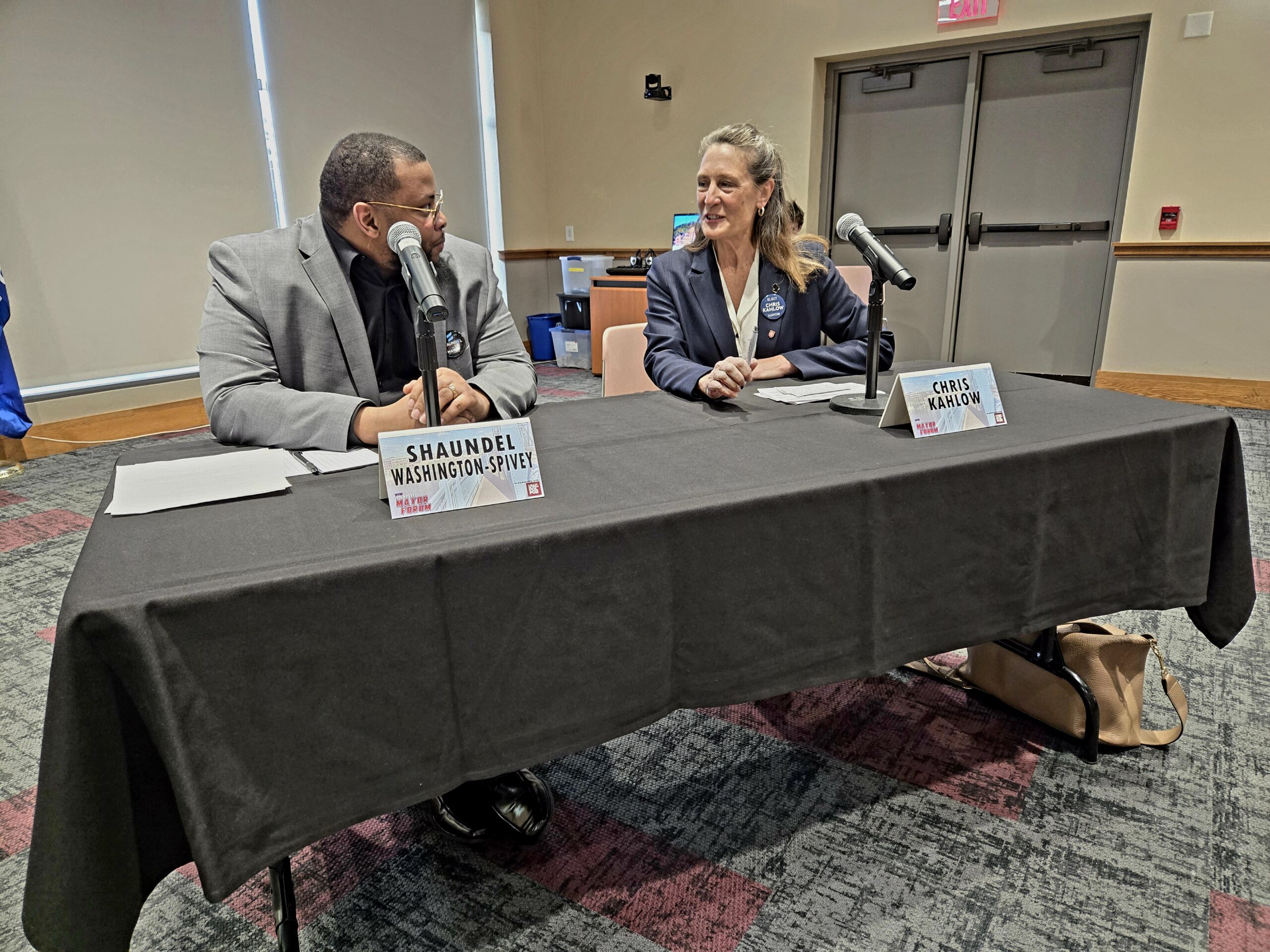 Two people sitting at a table with microphones, name tags read Shaundel Washington-Spivey and Chris Kulow. Theyre in a conference room with closed doors in the background.