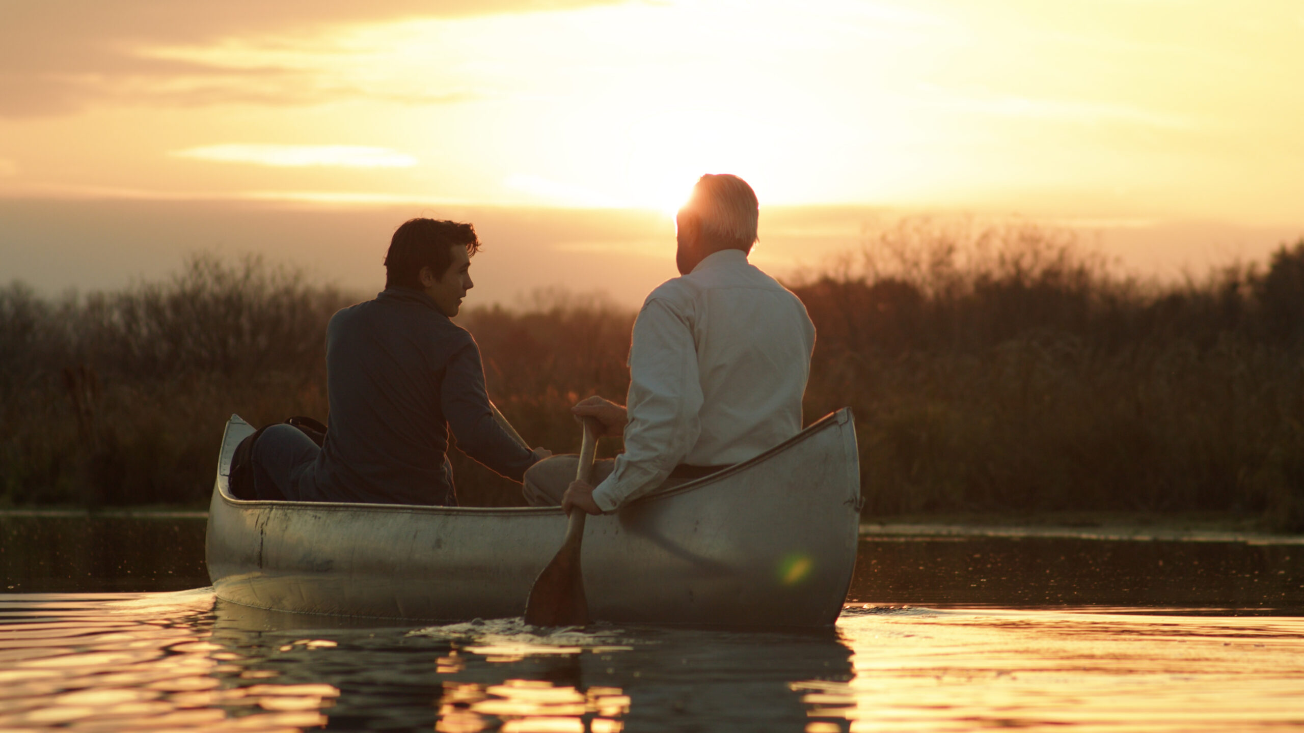 Two people in a canoe on a calm lake during sunset, with trees visible in the background.