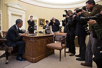 A person sits at a desk signing papers while a group of photographers takes pictures in an office with striped walls and framed portraits.