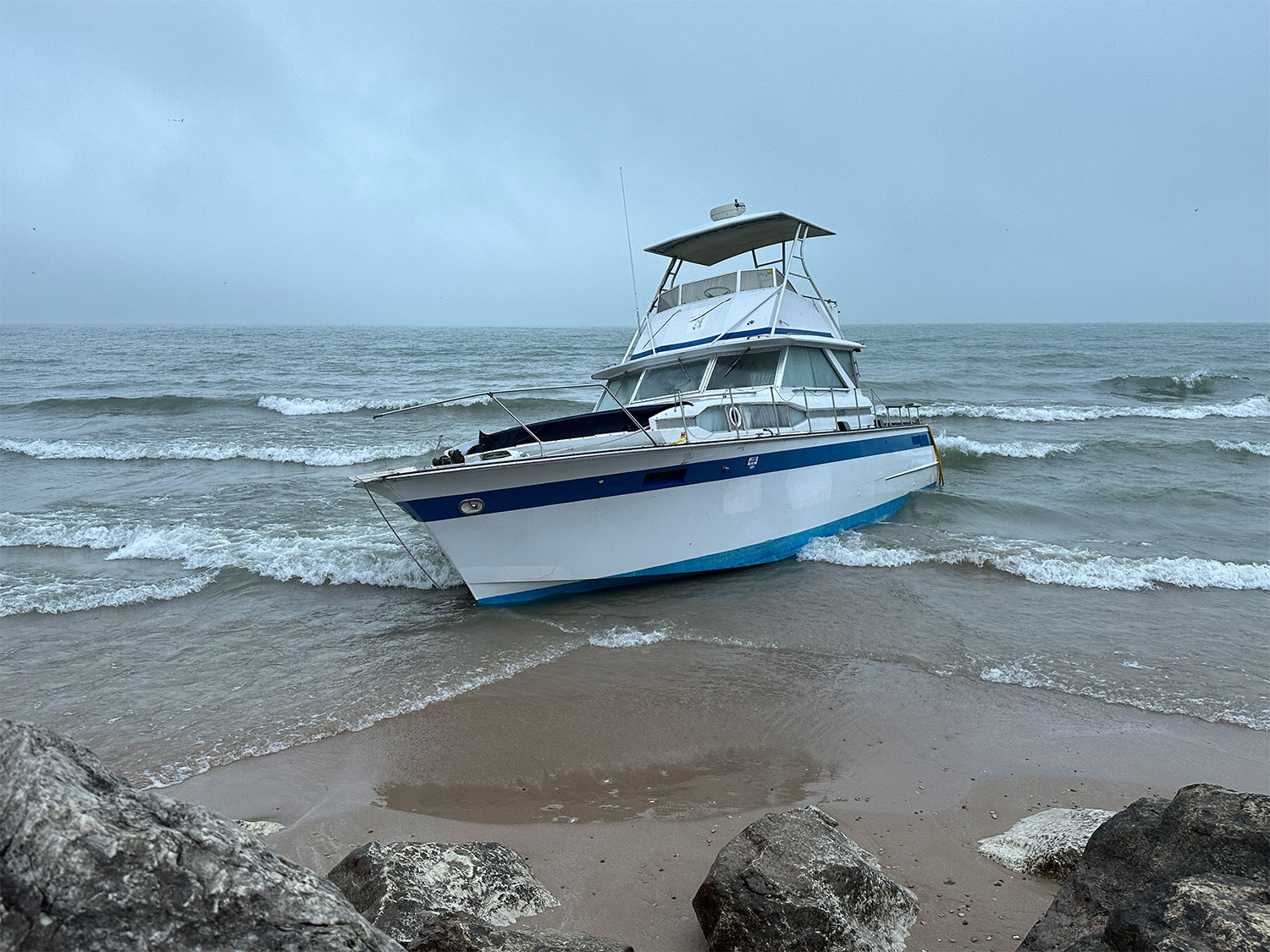 A blue and white boat is grounded on a sandy beach with rocky outcrops, under overcast skies and gentle waves.