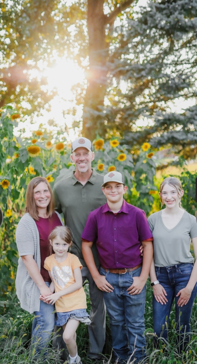 Family of five posing in front of sunflowers and trees, with sunlight filtering through.