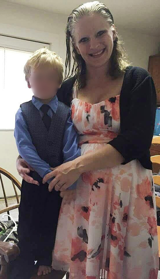 A woman in a floral dress poses indoors with a young boy in a blue shirt and vest. The boys face is blurred. They are standing in a domestic setting with a wooden chair behind them.