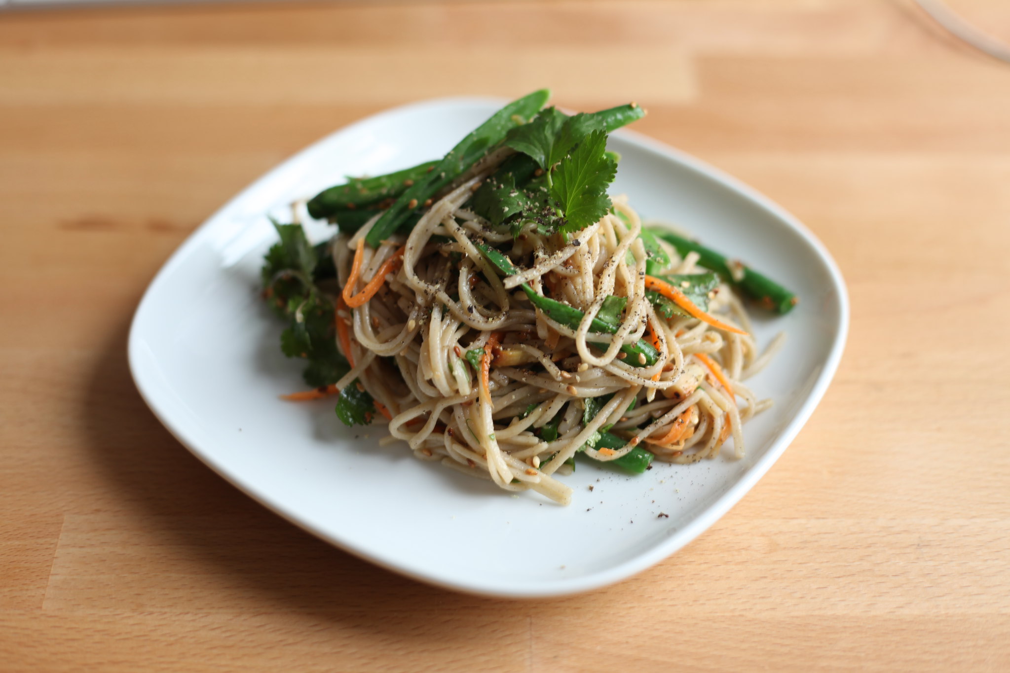 A plate of spaghetti with vegetables, garnished with greens and seasoned with pepper, sits on a wooden table.