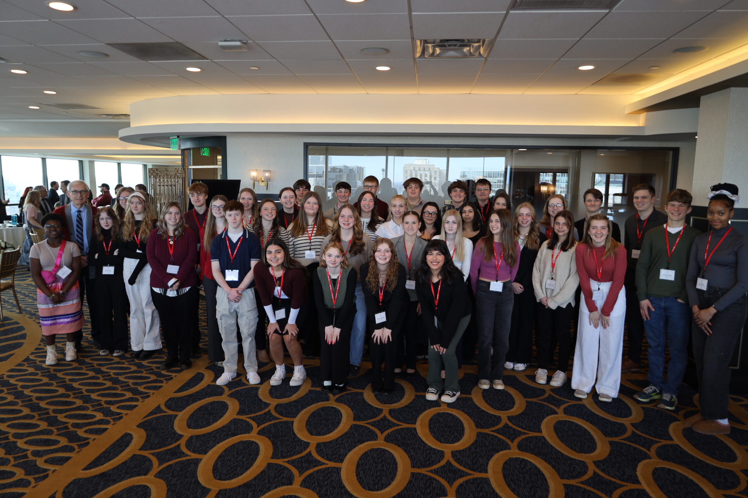 A large group of people, mostly young adults, stand together in a conference room with patterned carpet and ceiling lights, wearing name tags and red lanyards.