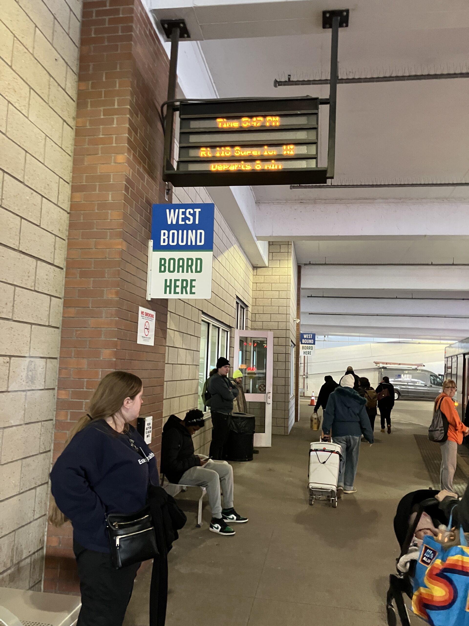 People wait at a bus stop with a digital display showing bus times. A sign reads West Bound Board Here. Some individuals are seated while others stand.