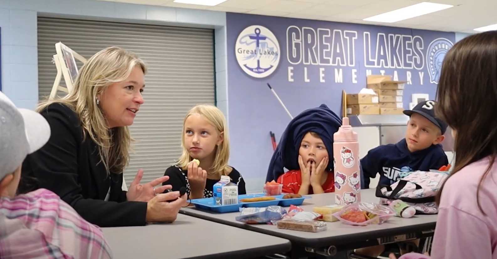 A woman speaks with four children seated at a lunch table in a school cafeteria, with a wall displaying Great Lakes Elementary in the background.