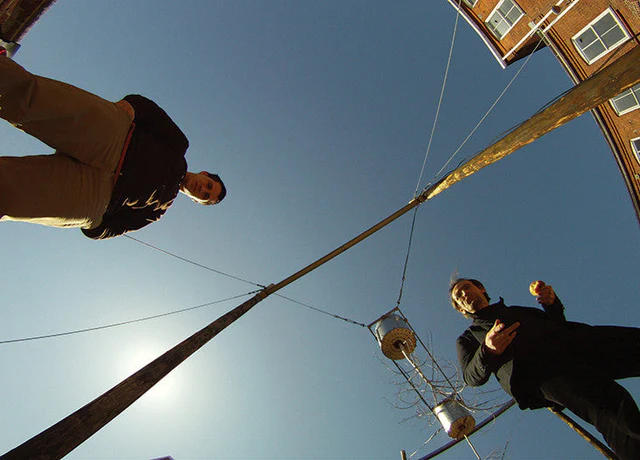 Two people are standing on either side of a street lamp pole, viewed from below. A clear blue sky and part of a brick building are visible in the background.