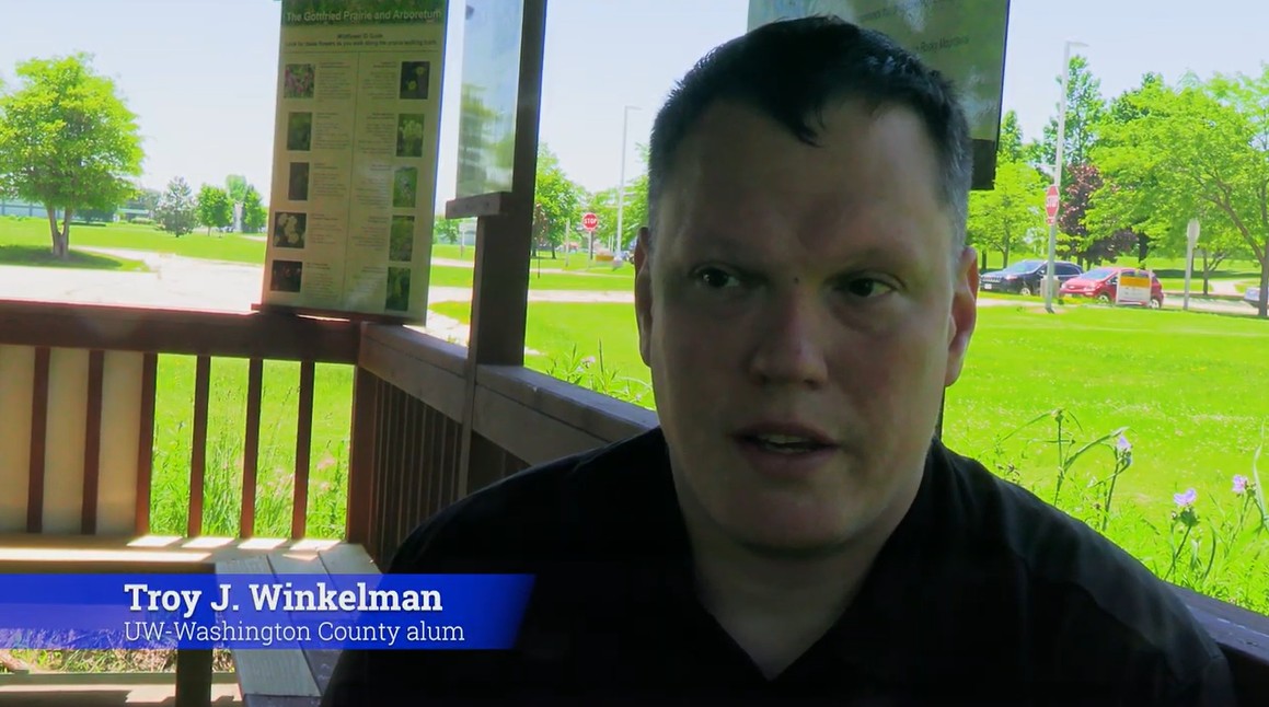 A man sits under a pavilion with a park in the background. Text on screen reads Troy J. Winkelman, UW-Washington County alum.