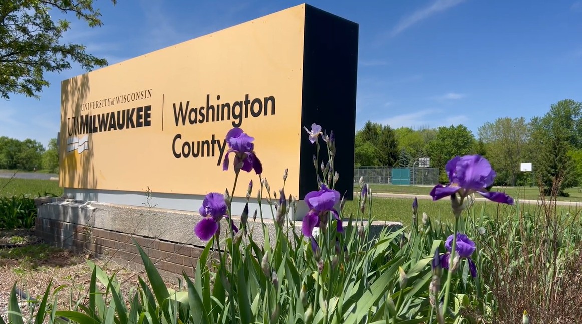 Sign for University of Wisconsin-Milwaukee, Washington County, with purple flowers in front. Clear blue sky in the background.