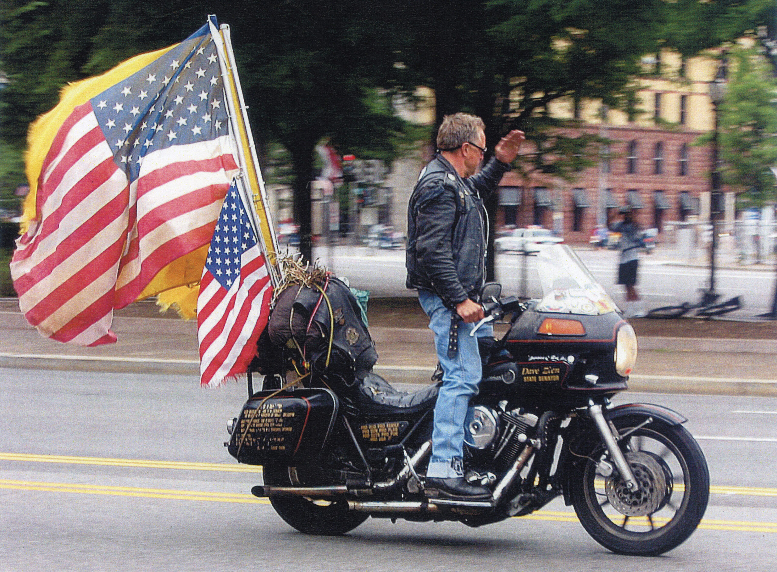 A person rides a motorcycle on a street, flying two large American flags behind.