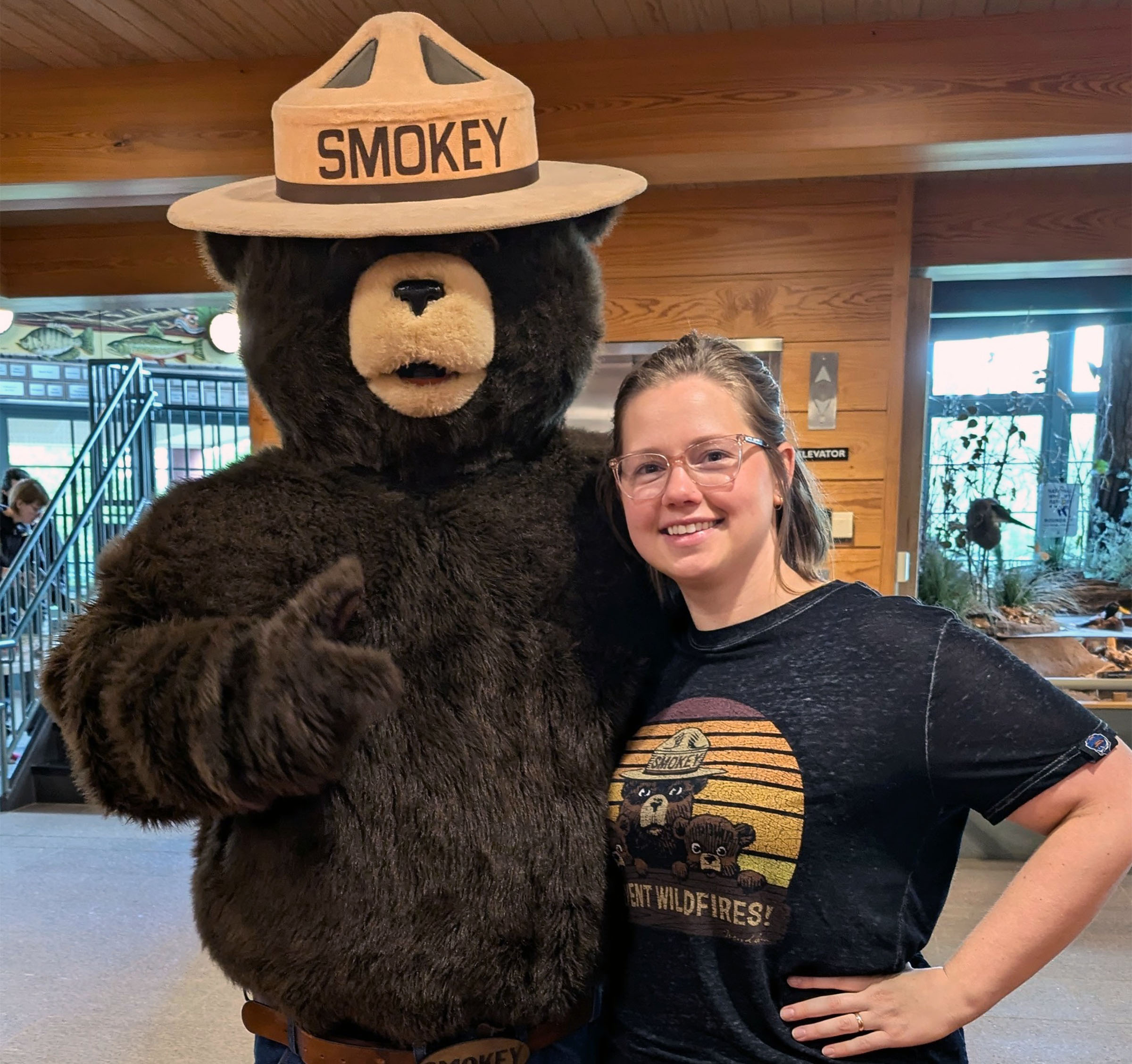 A person stands next to a person in a Smokey Bear costume indoors.