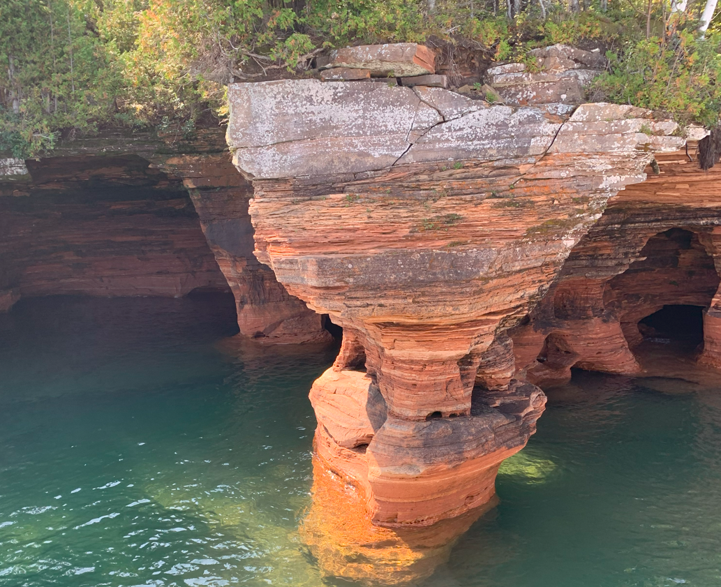 Rock formation with layered, weathered red rock above green water, surrounded by foliage.