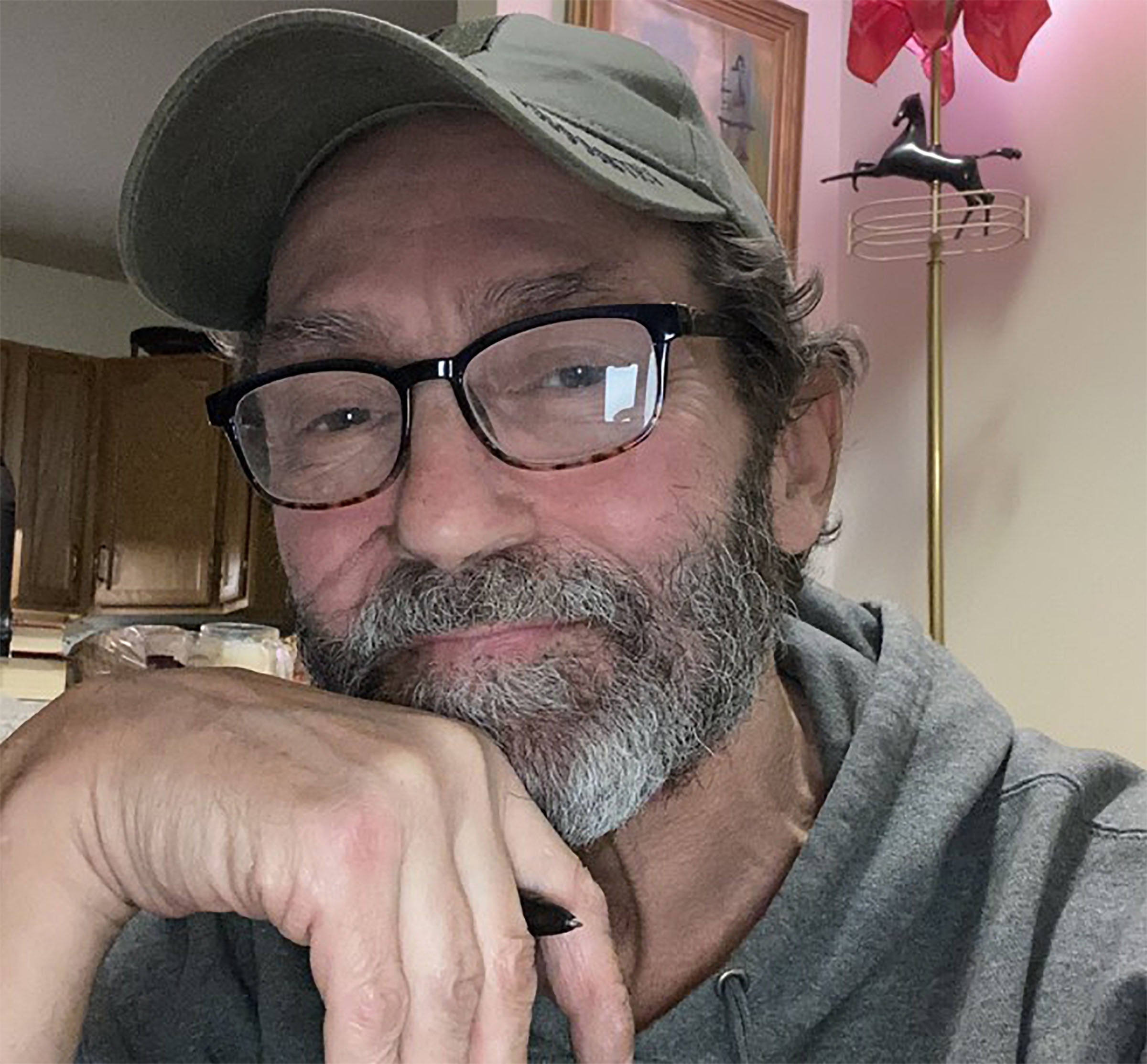 Man with glasses and a beard wearing a cap and hoodie, sitting indoors.